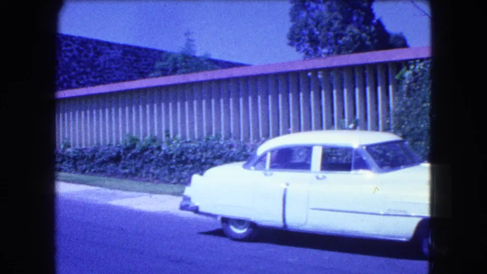 1962 Mexico City frame showing the rear and side of a white sedan near a curb, with hedges, a slatted fence, and bright 8mm color shift.