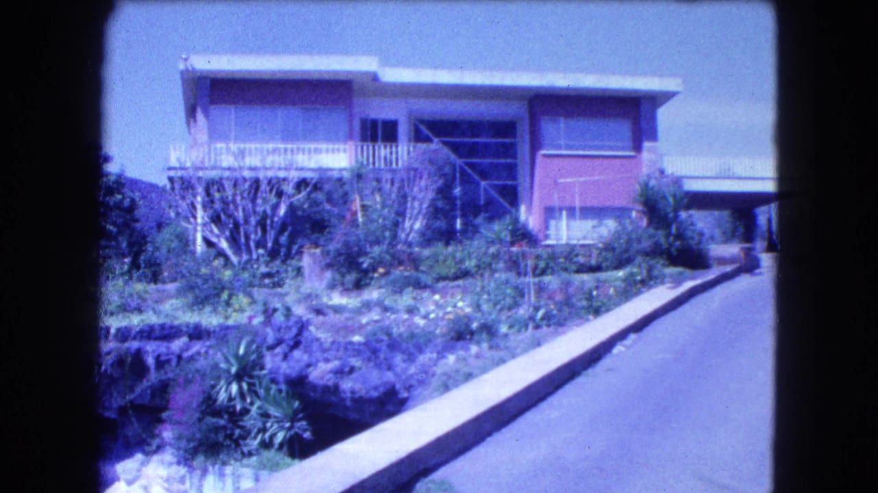 1962 Mexico City home-movie frame showing a modernist house with balcony, landscaped front garden, and paved driveway.