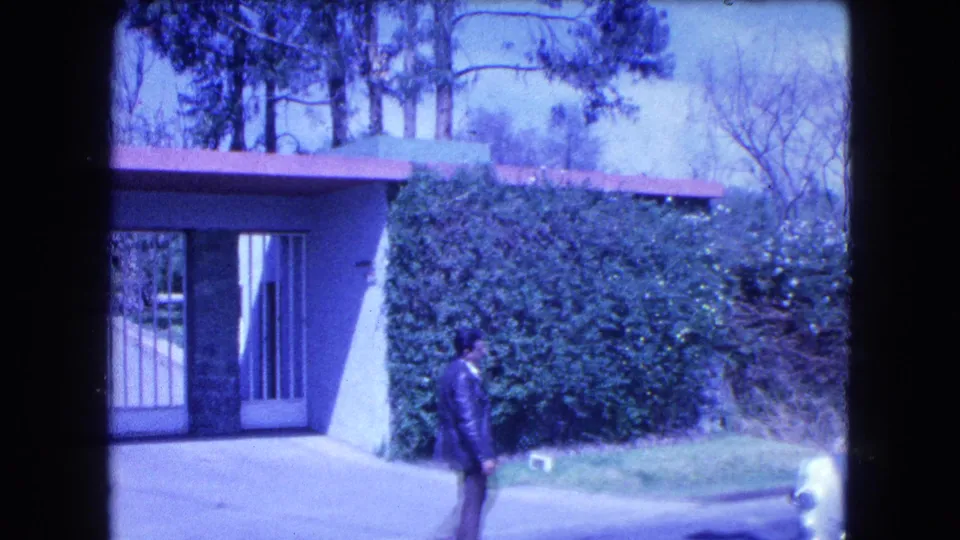 1962 Mexico City frame showing a gated mansion entrance, tall hedge, driveway, and an adult man in a dark suit.