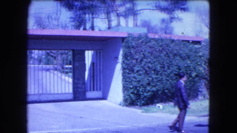 1962 Mexico City home-movie frame with a suited adult man walking near a gated residential entrance and hedge.