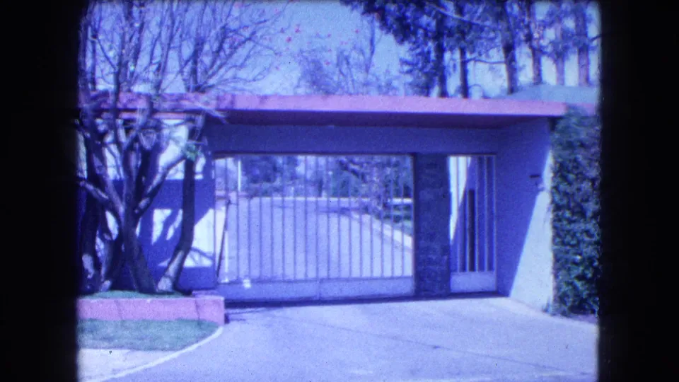 1962 Mexico City frame showing the mansion's closed driveway gate, adjacent hedge, tree, low wall, and entrance structure.