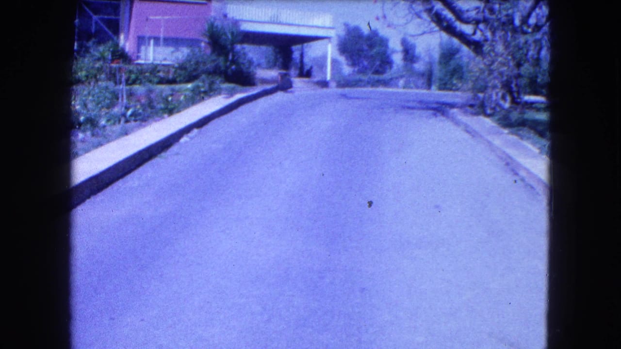 1962 Mexico City 8mm frame showing a paved residential road approaching a two-story house with shrubs and a garden edge.