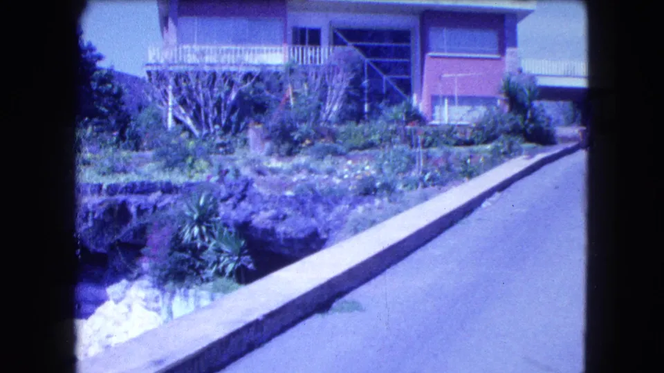 1962 home-movie frame showing landscaped plants, a concrete curb, and a modern residential house in Mexico City.