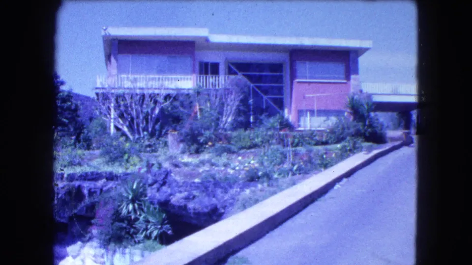 Blue-tinted 8mm frame of a Mexico City house with balcony, large windows, garden shrubs, and a roadside curb.