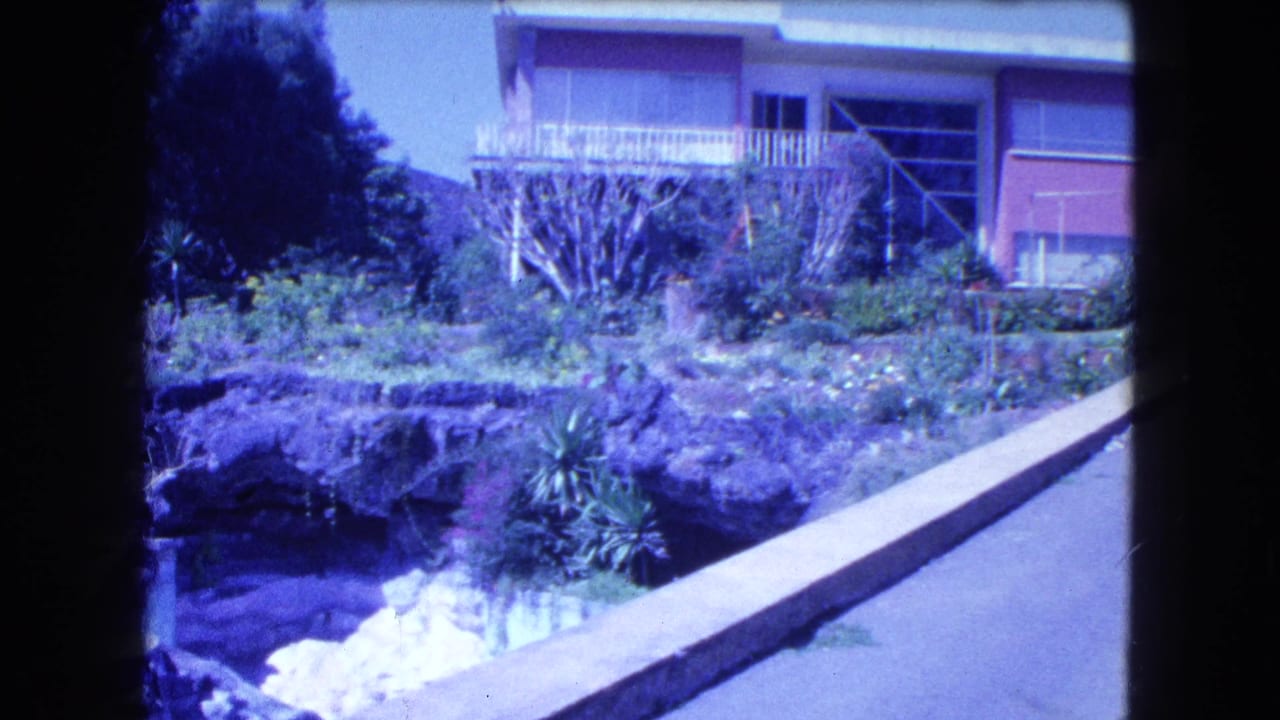1962 Mexico City home-movie frame showing a modern hillside house, balcony, garden, rock edging, and sloped driveway.