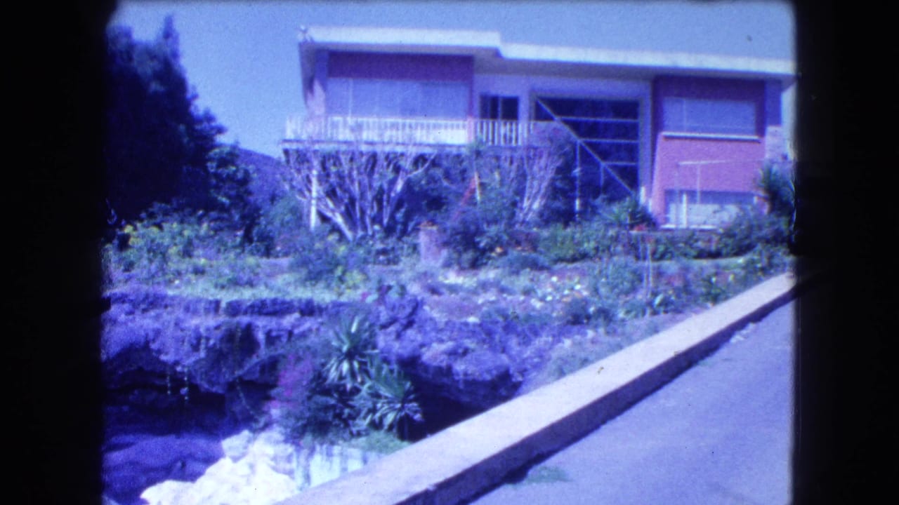 House exterior with balcony, garden plantings, rock edge, and driveway on a sunny slope.