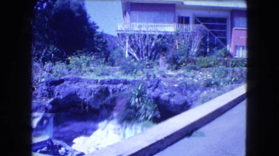 1962 Mexico City home-movie frame with terraced garden, reflective pool, shrubs, diagonal wall, and a modern house balcony.