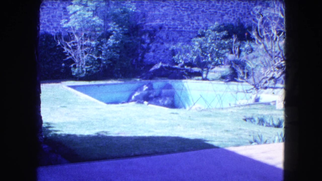 1962 Mexico City frame showing landscaped grounds, a greenhouse-like structure, lawn, shrubs, trees, and a high stone wall.
