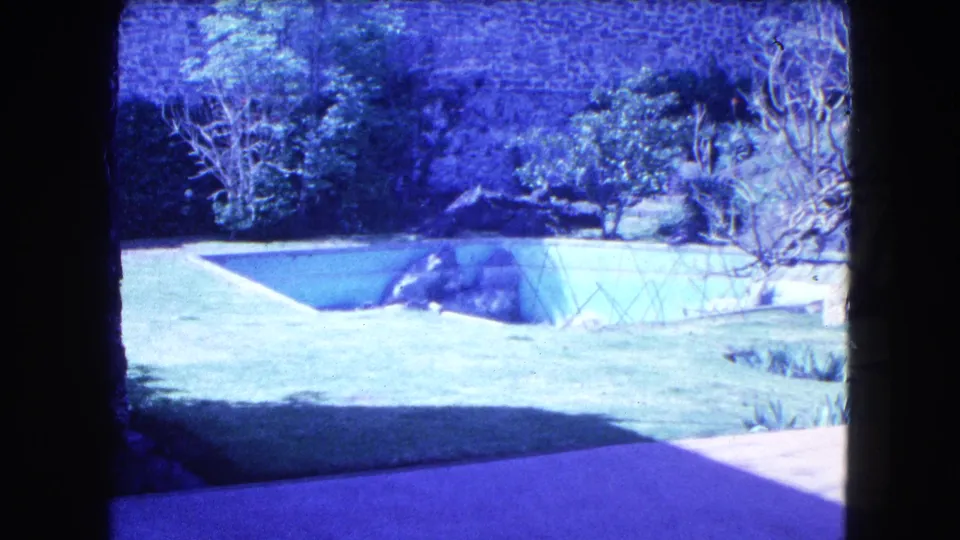 1962 Mexico City frame showing landscaped grounds, a greenhouse-like structure, lawn, shrubs, trees, and a high stone wall.