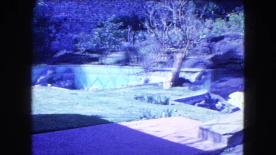1962 Mexico City frame showing a sunlit landscaped area with greenhouse-like structure, lawn, terrace, trees, and garden beds.