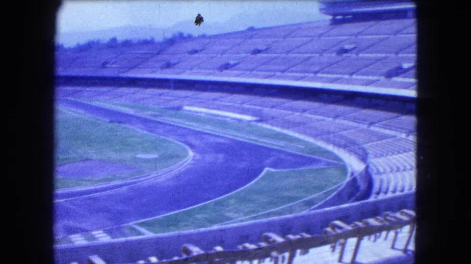 Elevated 1962 Mexico City view across an empty arena, showing concrete tiers, track lanes, field grass, and distant haze.