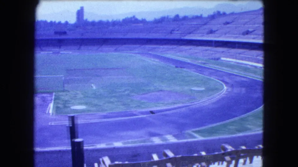 1962 Mexico City stadium frame with an empty grass field, track, goal frames, concrete stands, and hazy distant structures.