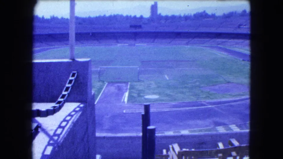 Home-movie frame from Mexico City in 1962 showing the empty stadium field, track, goal frame, seating, and a foreground pole.