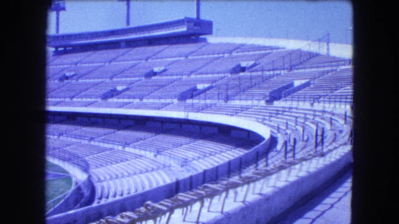 1962 Mexico City 8mm frame showing empty curved stadium stands, railings, concrete barriers, and a partial green field.