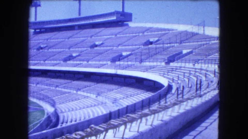 1962 Mexico City 8mm frame showing empty curved stadium stands, railings, concrete barriers, and a partial green field.