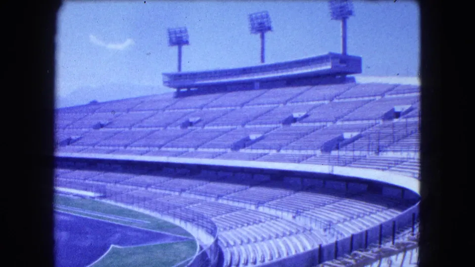 1962 Mexico City 8mm frame showing empty stadium tiers, floodlight towers, railings, and a partial green field below.