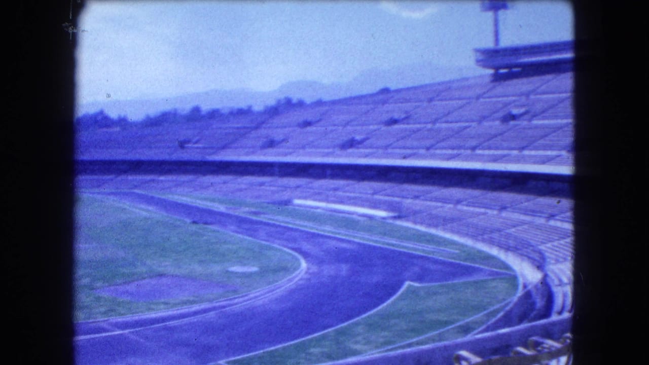1962 Mexico City 8mm frame showing empty grandstands, a curved running track, field edges, and distant mountains.