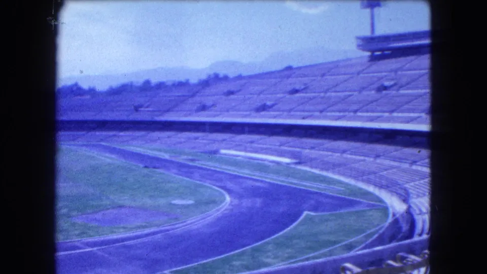 1962 Mexico City 8mm frame showing empty grandstands, a curved running track, field edges, and distant mountains.