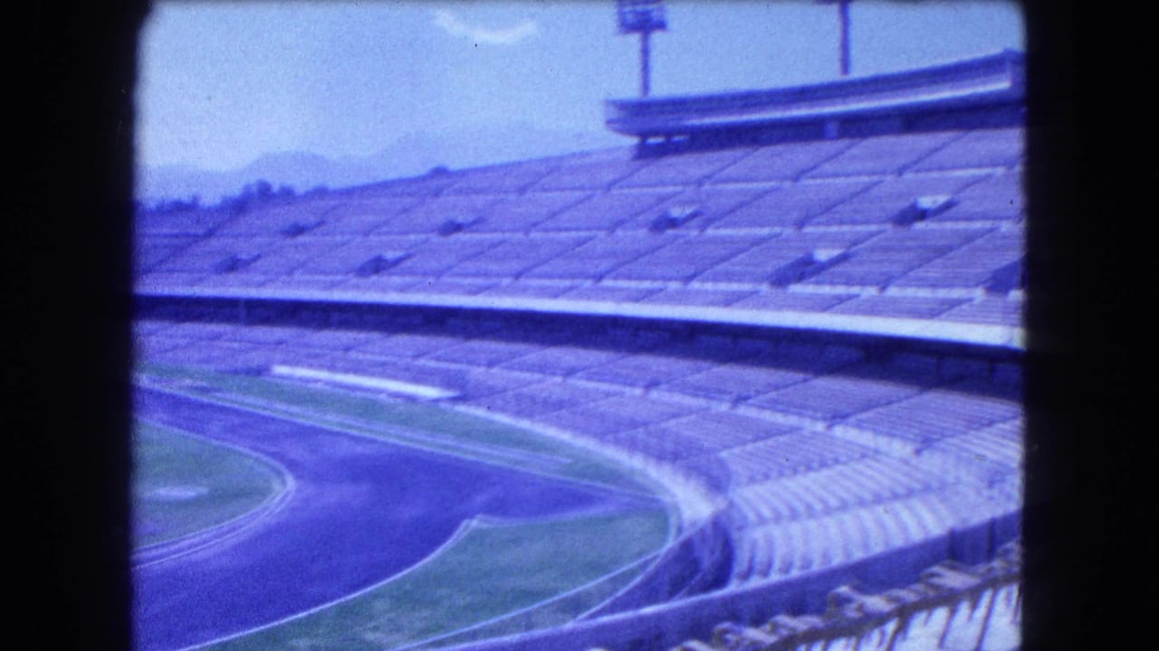 1962 Mexico City stadium view with tiered seating, a blue-toned running track, floodlight towers, and mountains beyond the venue.