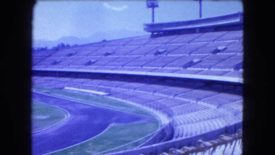 1962 Mexico City stadium view with tiered seating, a blue-toned running track, floodlight towers, and mountains beyond the venue.