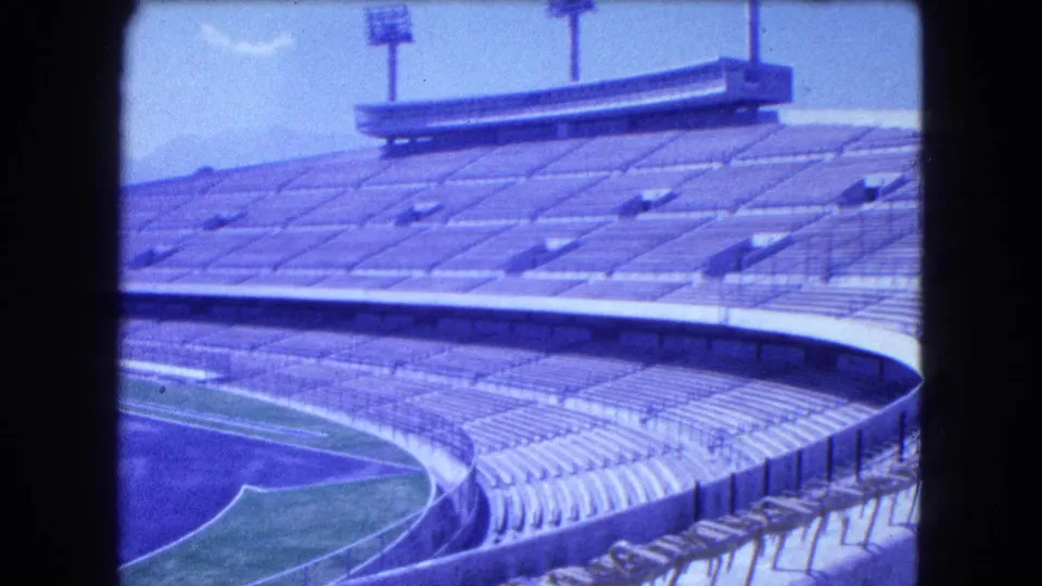 1962 Mexico City 8mm frame of empty concrete stands, a curved upper rim, floodlight poles, and a large structure above the seating.