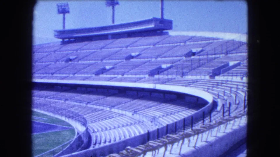 1962 Mexico City frame showing the stadium's empty grandstands, curved track edge, railings, floodlight tower, and bright sky.