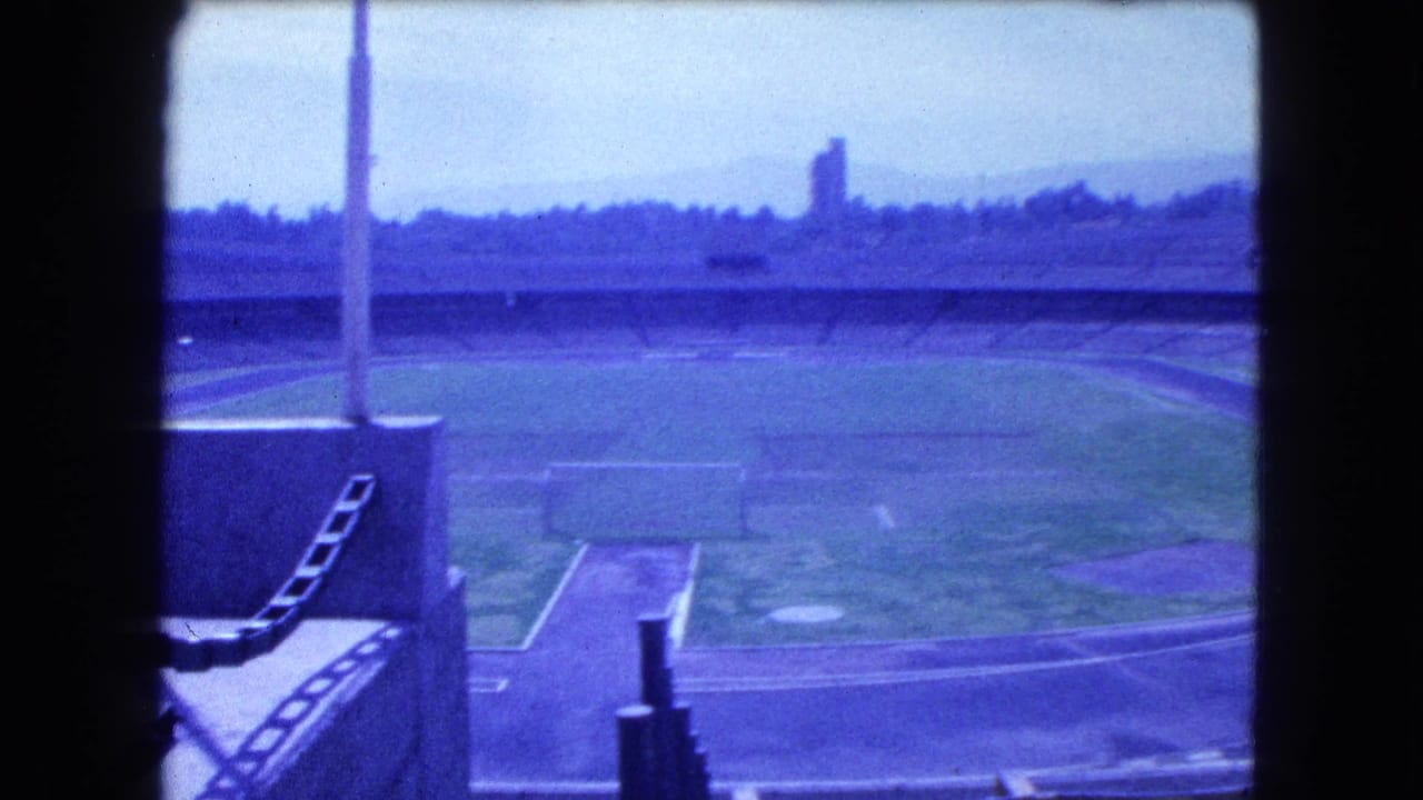 Wide 8mm frame of an empty Mexico City stadium in 1962, showing the oval track, infield, tiered seating, and hazy horizon.