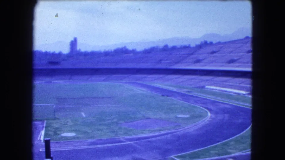 Empty 1962 Mexico City stadium view from an elevated visitor angle, with curved track, infield, concrete seating, and skyline haze.