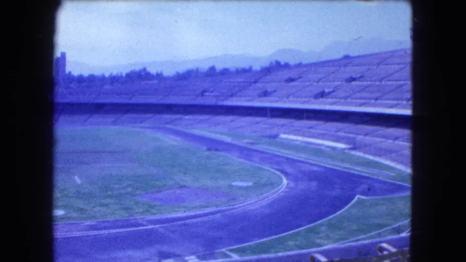 Blue-cast 8mm frame of an empty Mexico City track stadium in 1962, with concrete stands, curved lanes, field markings, and distant hills.