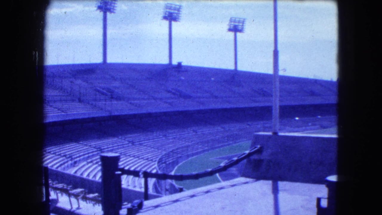 1962 Mexico City stadium view with blue-tinted empty seats, floodlight towers, railings, and dark 8mm frame edges.