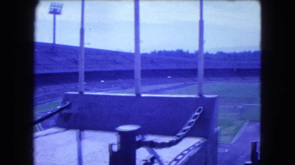 Elevated 1962 Mexico City stadium view with foreground posts, floodlight poles, empty seating, and a green playing surface.