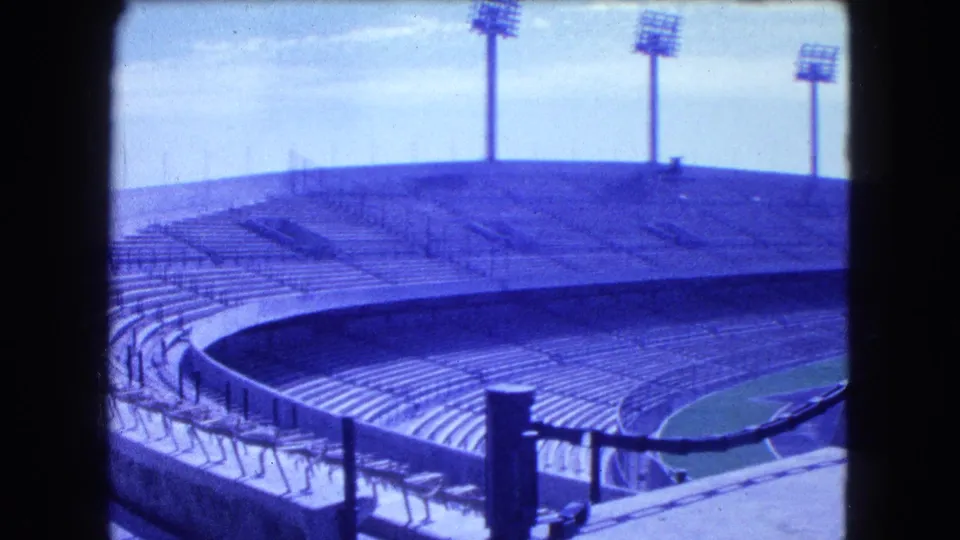 Visitor-shot 8mm frame of a large Mexico City stadium in 1962, with blue-tinted seating tiers, railings, and floodlight towers.