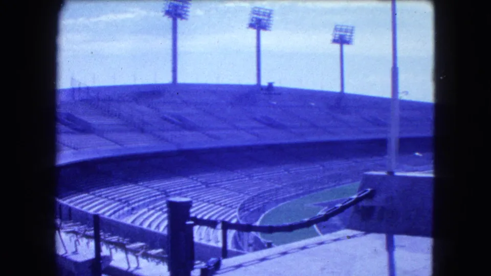 1962 8mm frame from Mexico City showing an empty stadium bowl, foreground railing hardware, concrete stands, and tall lights.