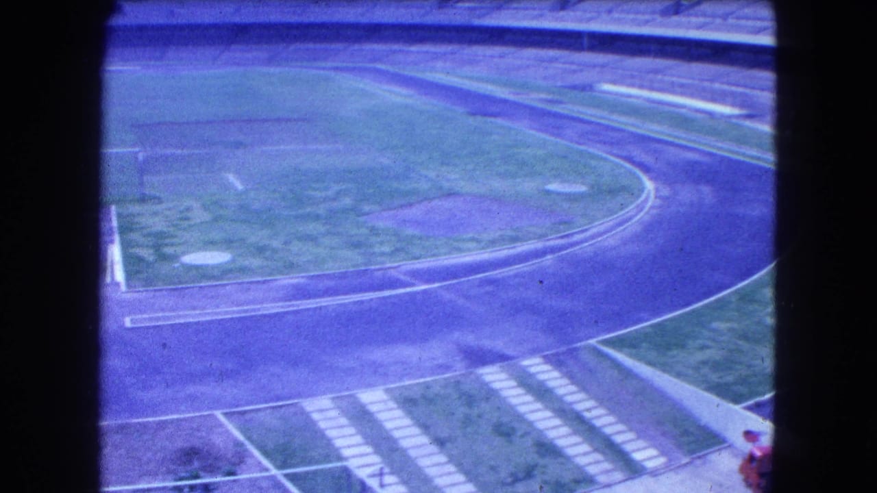 1962 Mexico City 8mm view of an empty stadium with a curved track, grass infield, and concrete bleachers under a blue-purple film cast.