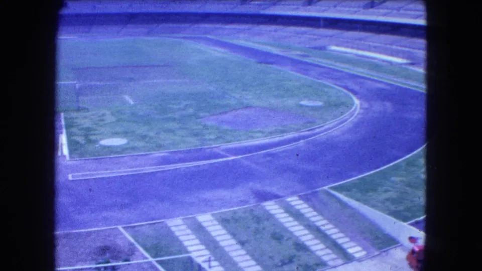 1962 Mexico City 8mm view of an empty stadium with a curved track, grass infield, and concrete bleachers under a blue-purple film cast.