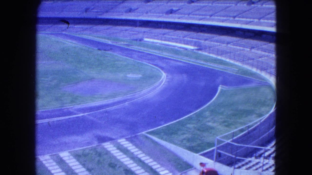 Empty stadium track curves past grass and seating, with a small distant person near the lower edge.