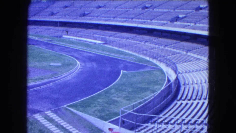 1962 Mexico City 8mm frame showing the stadium track bend, grassy infield, railings, and long rows of vacant concrete bleachers.