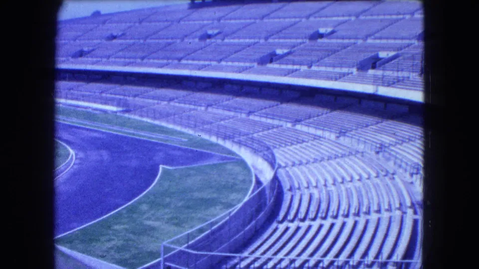 1962 Mexico City stadium frame with empty concrete seating, a curved running track, grass borders, railings, and strong blue-purple 8mm color shift.