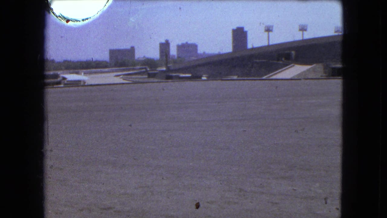 1962 Mexico City home-movie view across an open sports arena area toward empty stands and distant urban buildings.