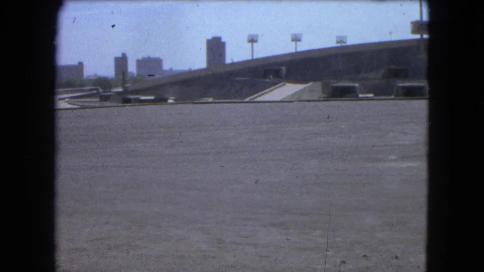 1962 Mexico City home-movie frame with a broad paved foreground, sloping concrete ramp, skyline buildings, and visible 8mm grain.