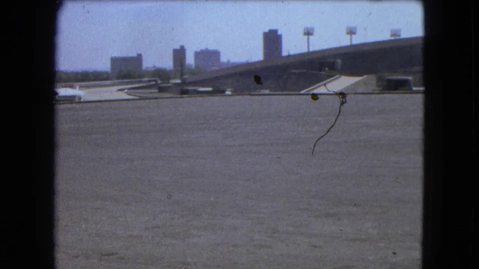 1962 Mexico City 8mm frame showing an elevated ramp, open paved ground, distant high-rises, light poles, and film dust marks.