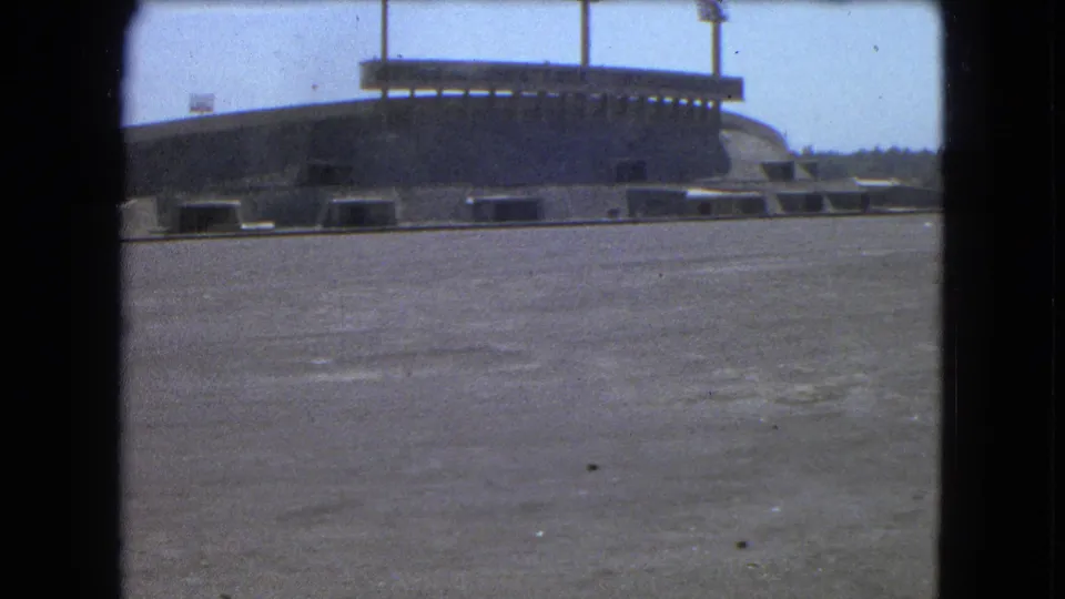 1962 Mexico City 8mm frame showing the stadium from a lower angle, with its curved structure, floodlights, and open pavement.