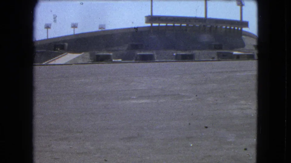1962 Mexico City frame showing an empty stadium exterior with lower openings, floodlight towers, and a broad concrete foreground.