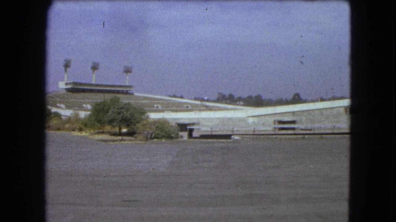 1962 Mexico City 8mm frame showing an empty concrete stadium, tall floodlight towers, small trees, and a wide paved area.
