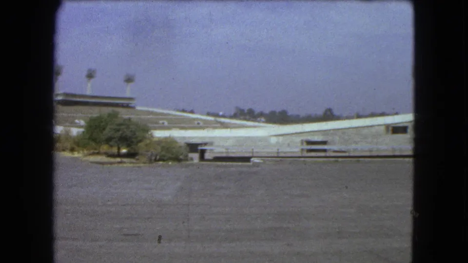 1962 Mexico City home-movie frame of a concrete stadium exterior with floodlight towers, shrubs, and open pavement.
