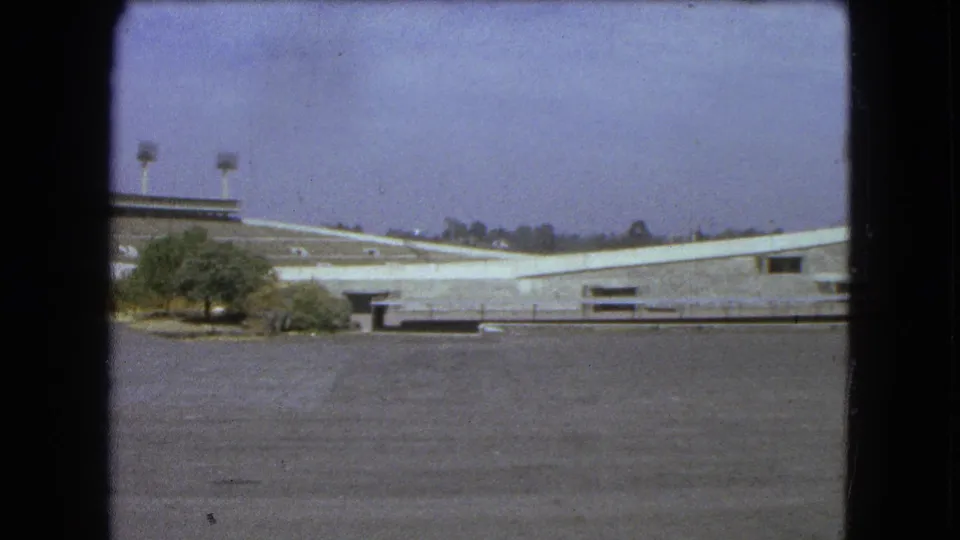 1962 Mexico City 8mm frame showing the stadium from a slightly lower position, with pavement, trees, and floodlights visible.