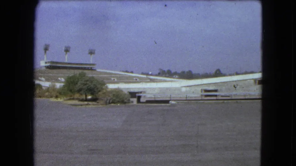 1962 Mexico City 8mm frame with a wide empty stadium view, foreground greenery, floodlight towers, and visible film wear.