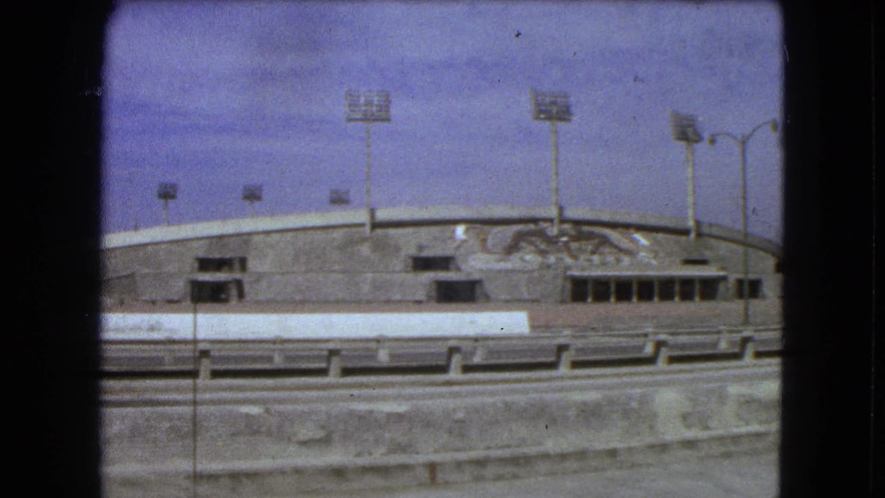 1962 Mexico City home-movie view of a large concrete stadium exterior with tall light towers and a mural on the facade.