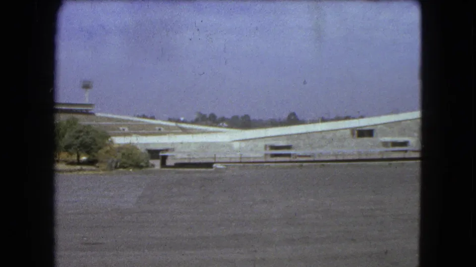 1962 Mexico City stadium exterior seen across an open paved forecourt with small trees at the left edge.