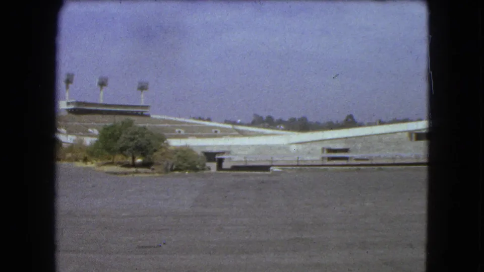 Faded 8mm frame from 1962 Mexico City showing the stadium roofline, light towers, small trees and open pavement.